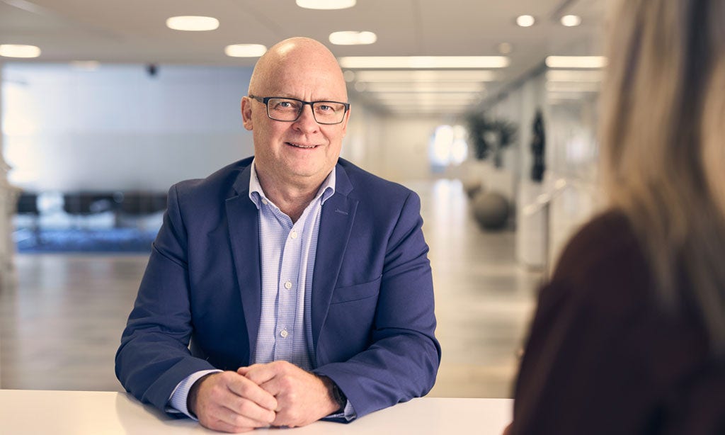 Man i blå kostym och glasögon, sitter vid skrivbord, ler och tittar in i kameran