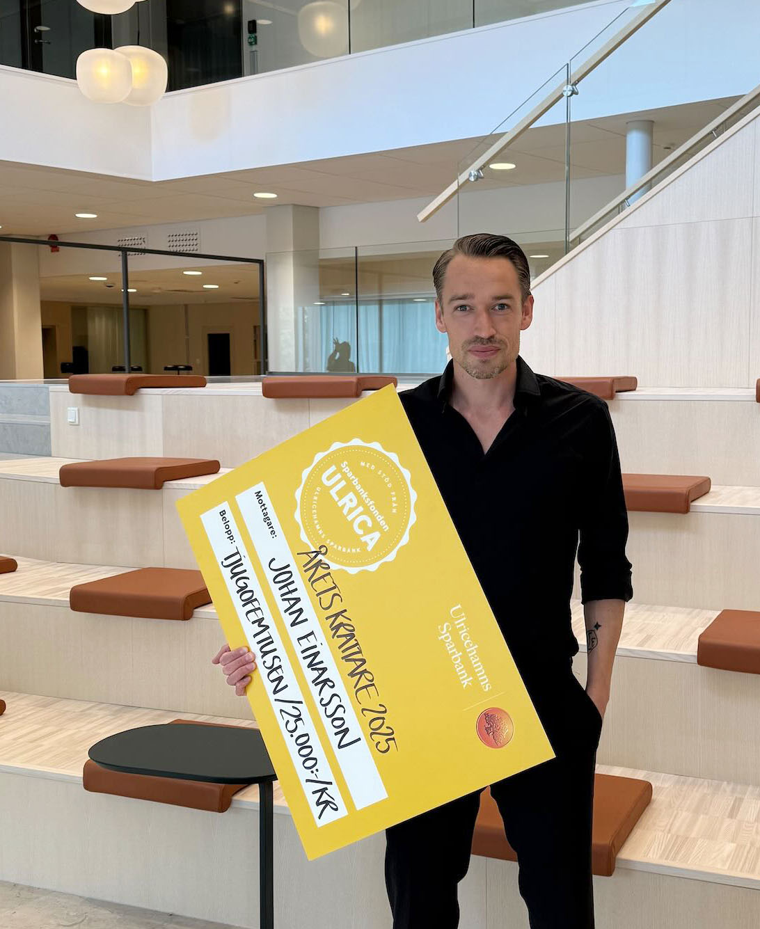 En man står i en gradängtrappa och håller i en gåvocheck med texten årets krattare 2025 - Johan Einarsson - 25 000 kr