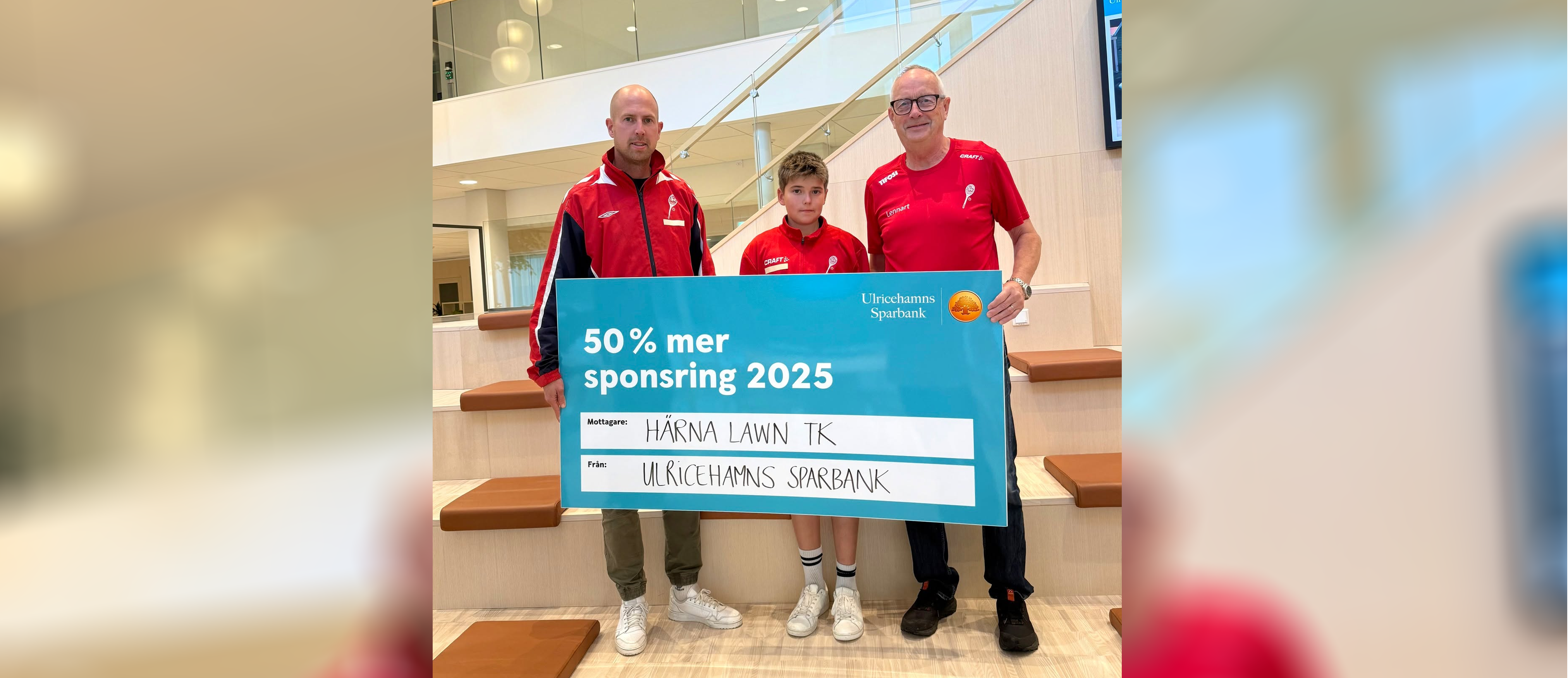 Två män och en ungdom i röda sportkläder står och håller i en turkost sponsringsscheck där det står 50 % mer ii sponsring till Härna Lawn TK