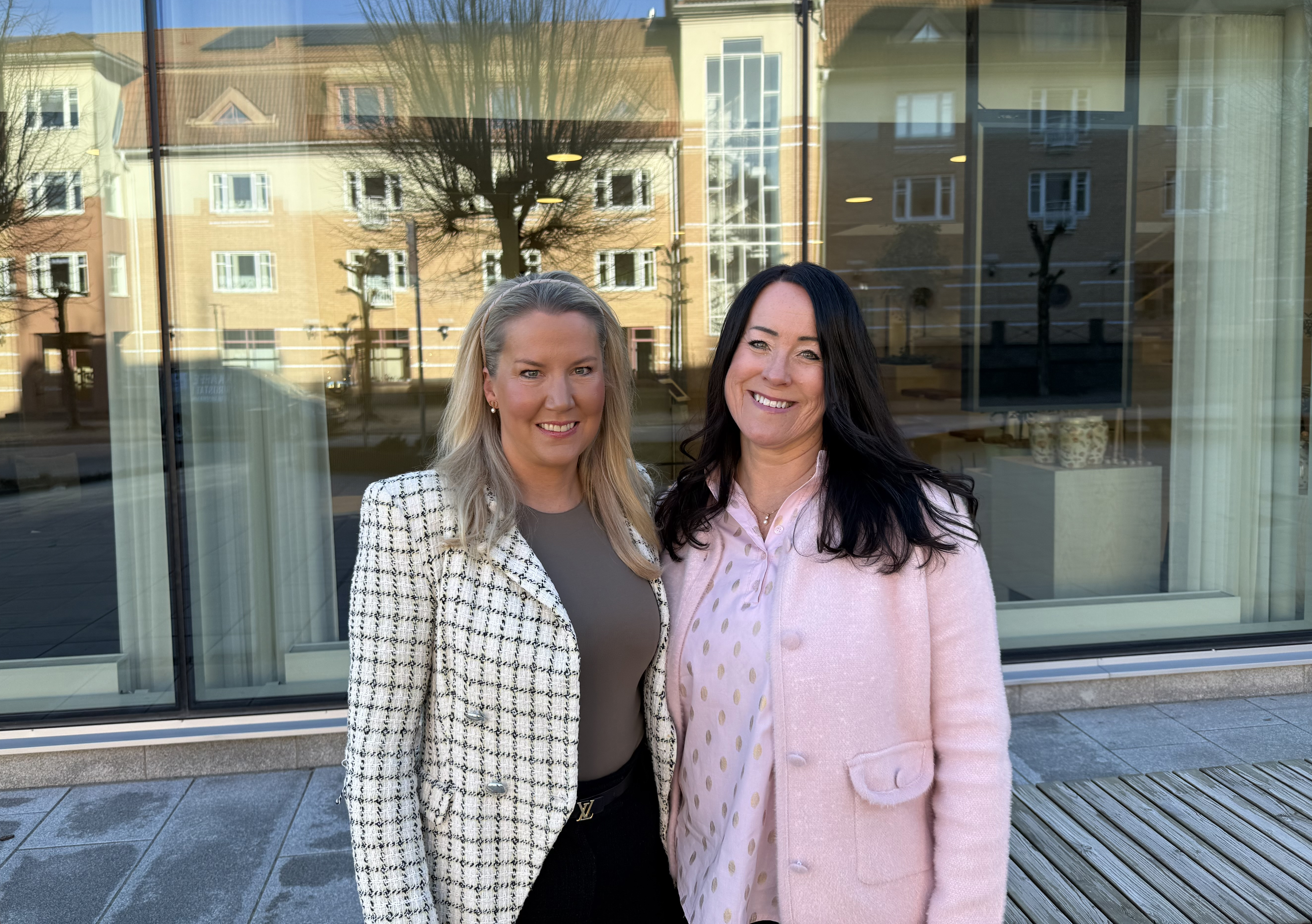 Två kvinnor står framför en bankbyggnad, tittar in i kameran och ler