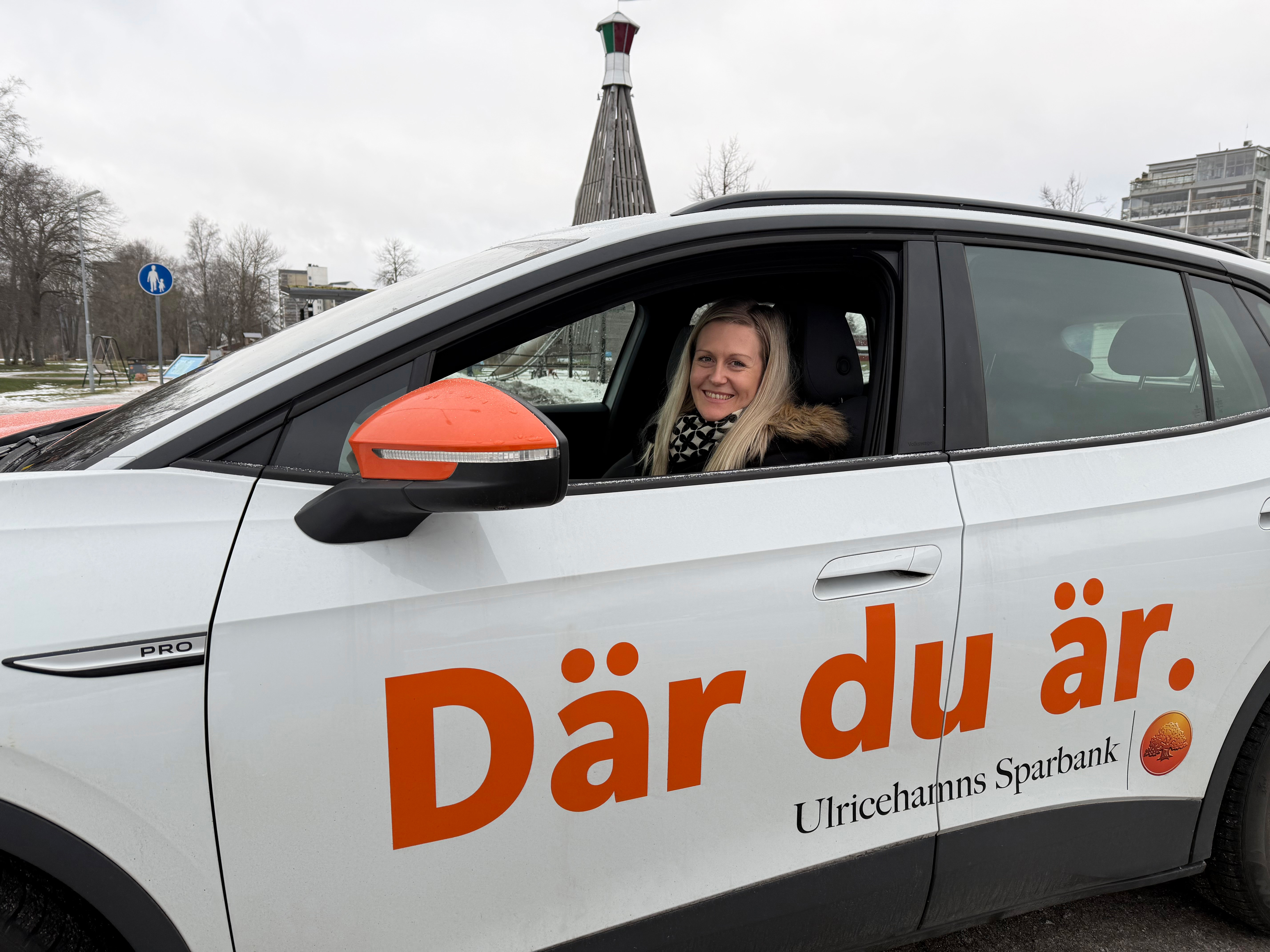 En kvinna sitter i förarsätet i en bil, rutan är nervevad, hon tittar in i kameran och ler