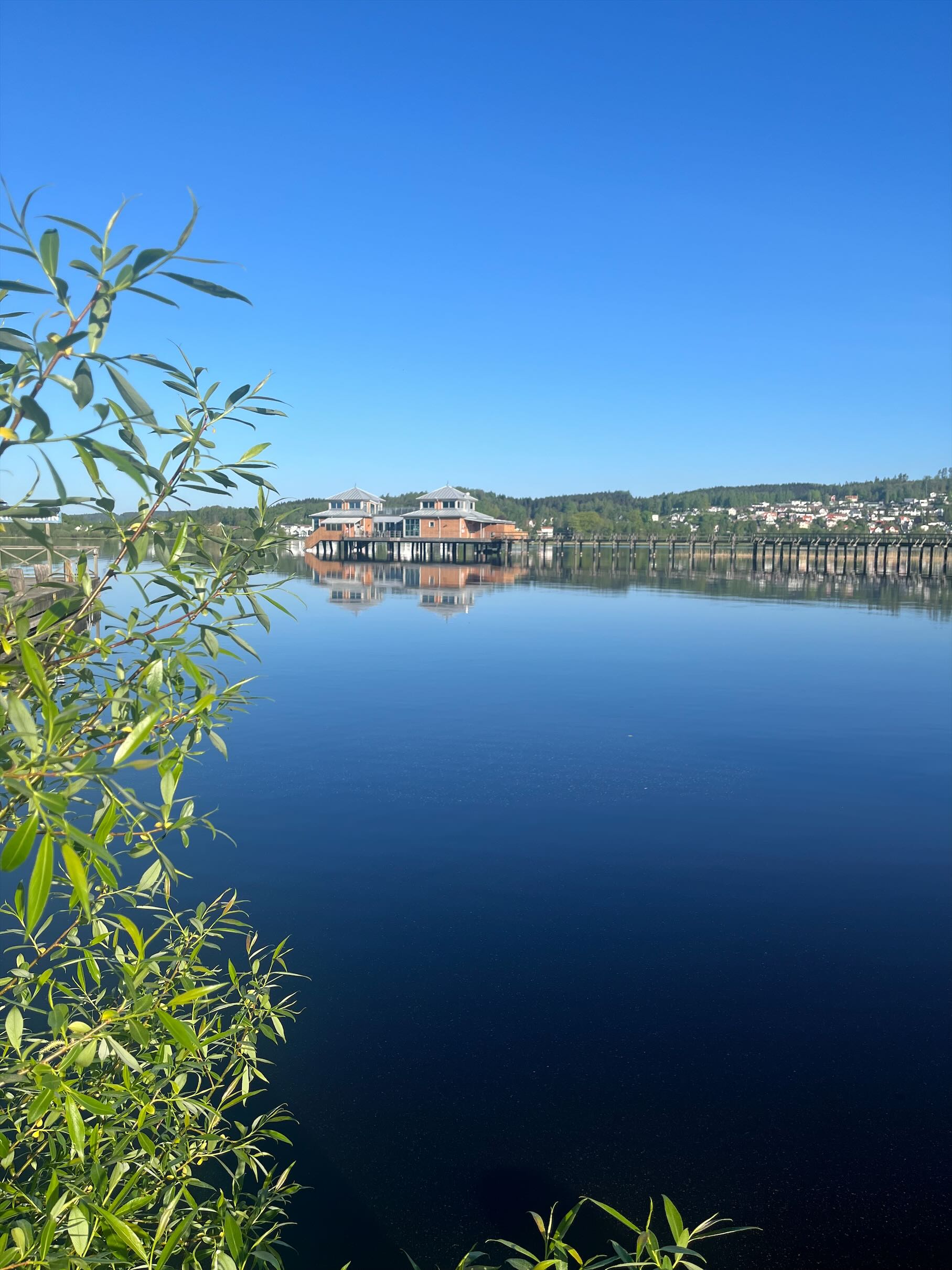En spegelblank blå sjö med blå himmel, lång brygga ut till ett kallbadhus