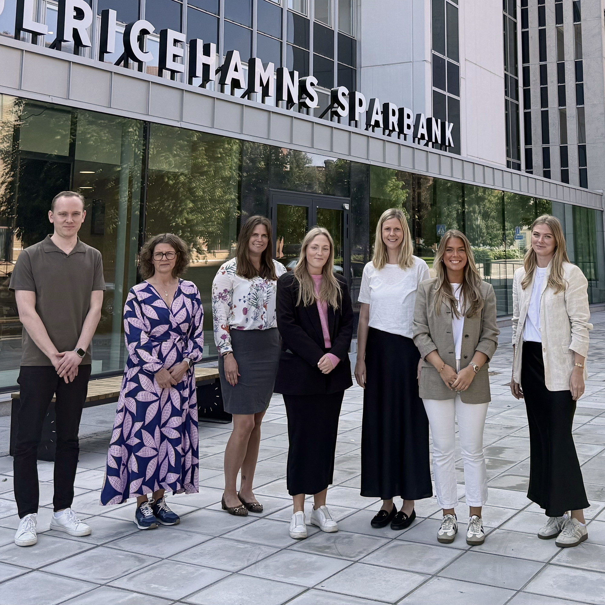 Några kvinnor och män står framför en bankentré, tittar in i kameran och ler