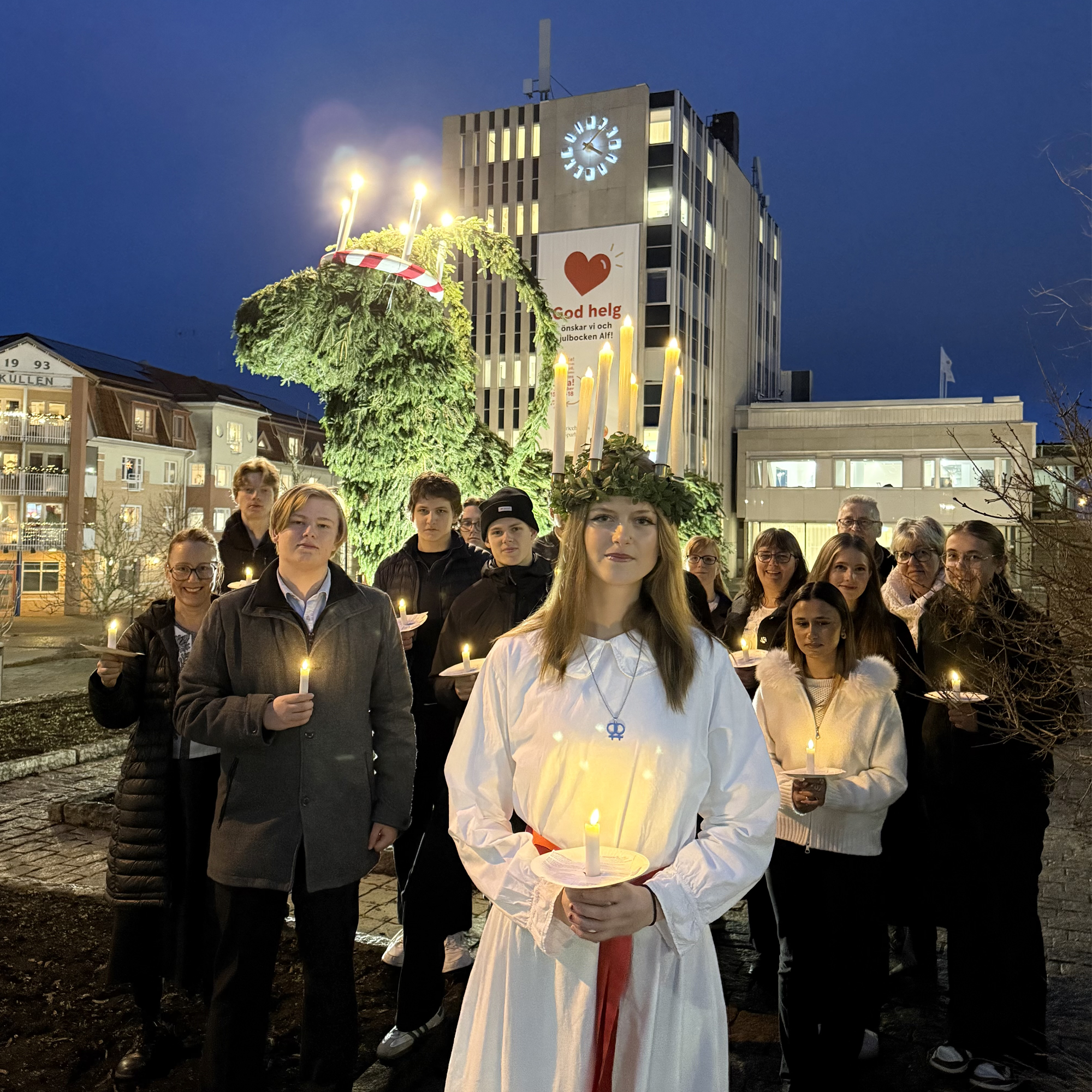 Ett luciatåg med ljus i händerna, längst fram står en lucia med en krona med ljus i, i förgrunden en stor glanbeklädd julbock med ljuskrona samt en bankbyggnad