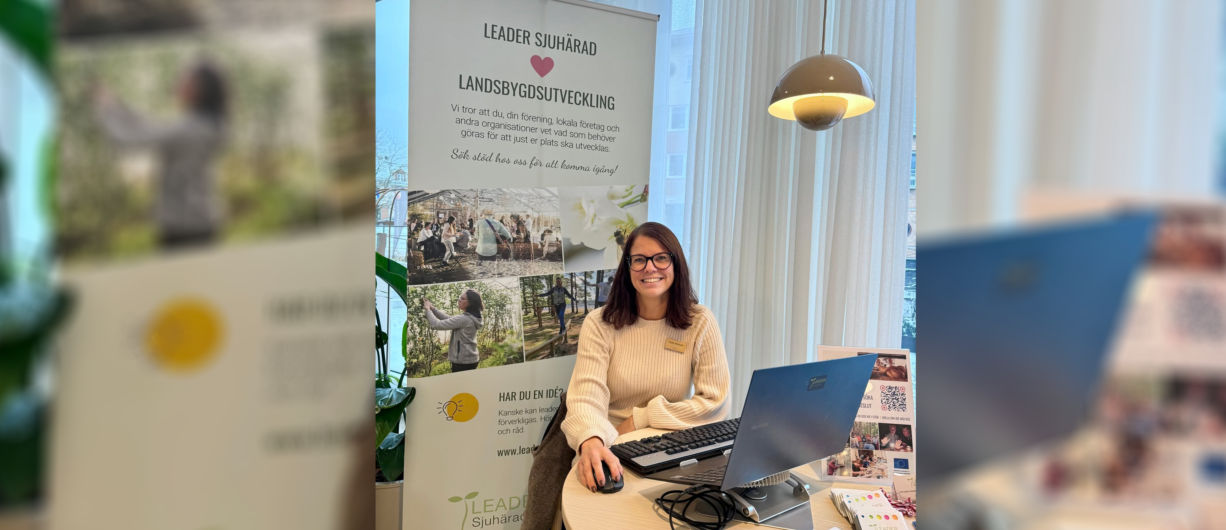 En kvinna sitter vid ett bord med en rollup bakom sig, hon tittar in i kameran och ler