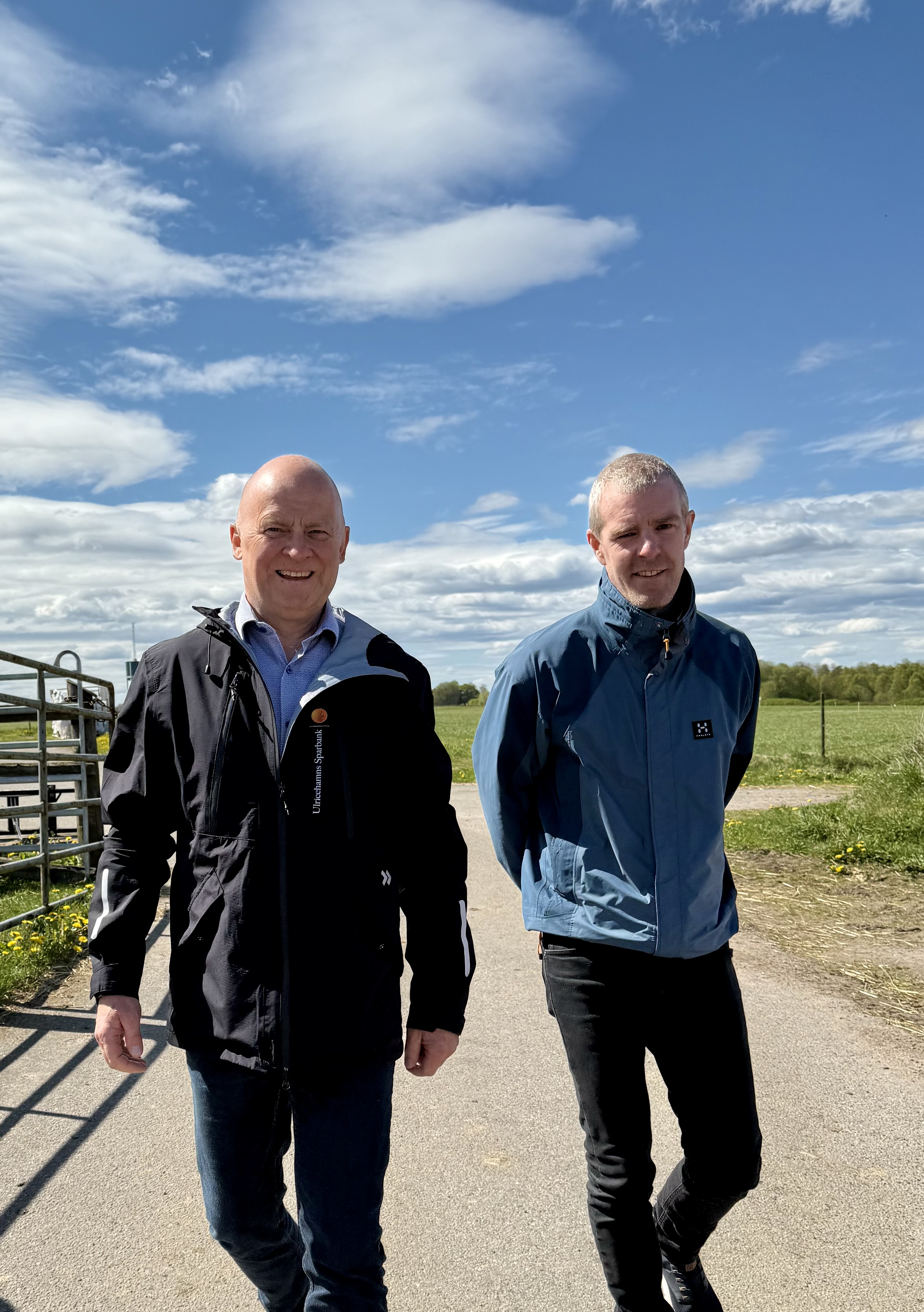 Två män går på en gård, blå himmel och sol, bägge tittar in i kameran och ler