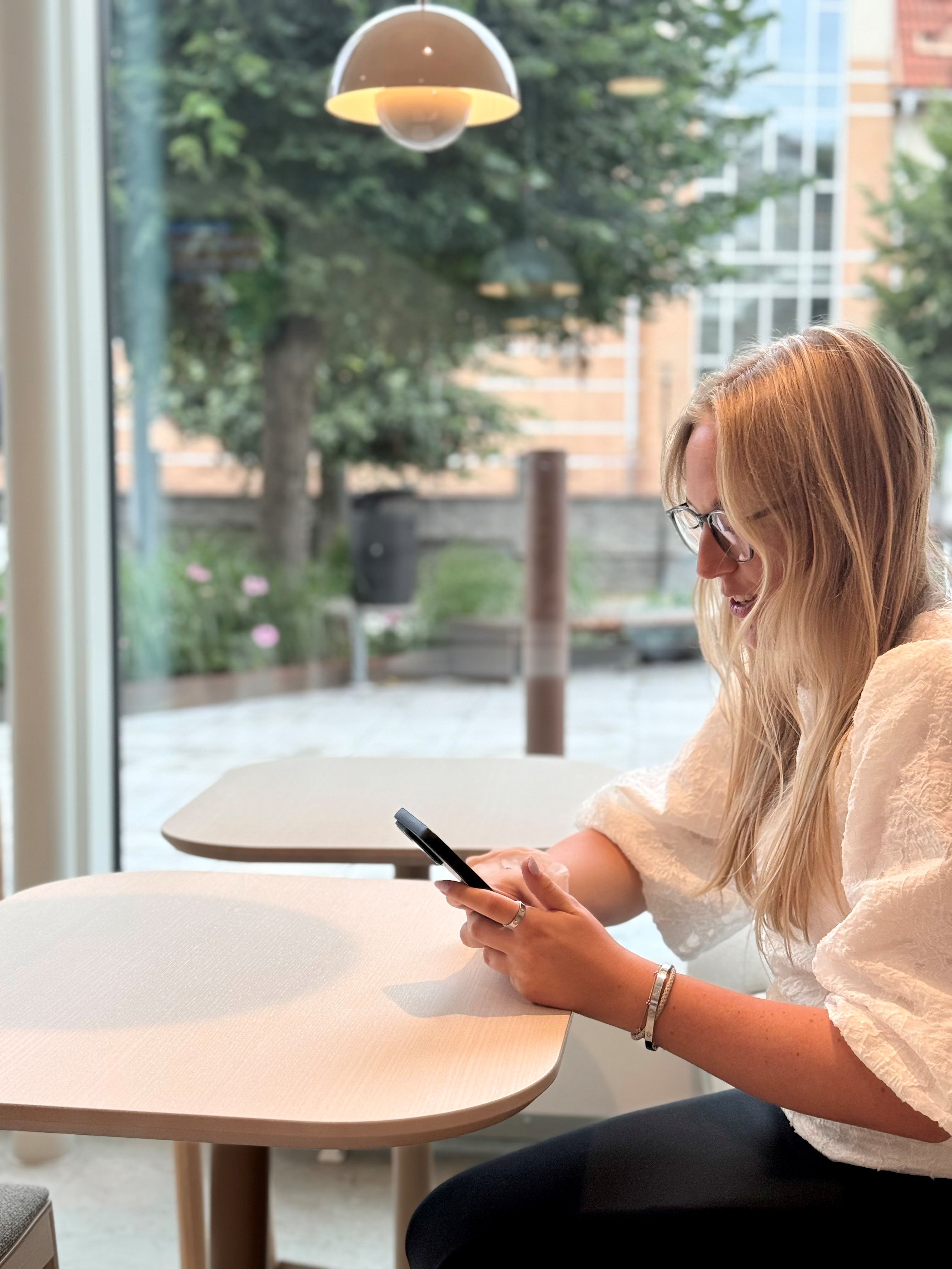 En kvinna sitter vid ett cafebord och tittar ner i en mobiltelefon