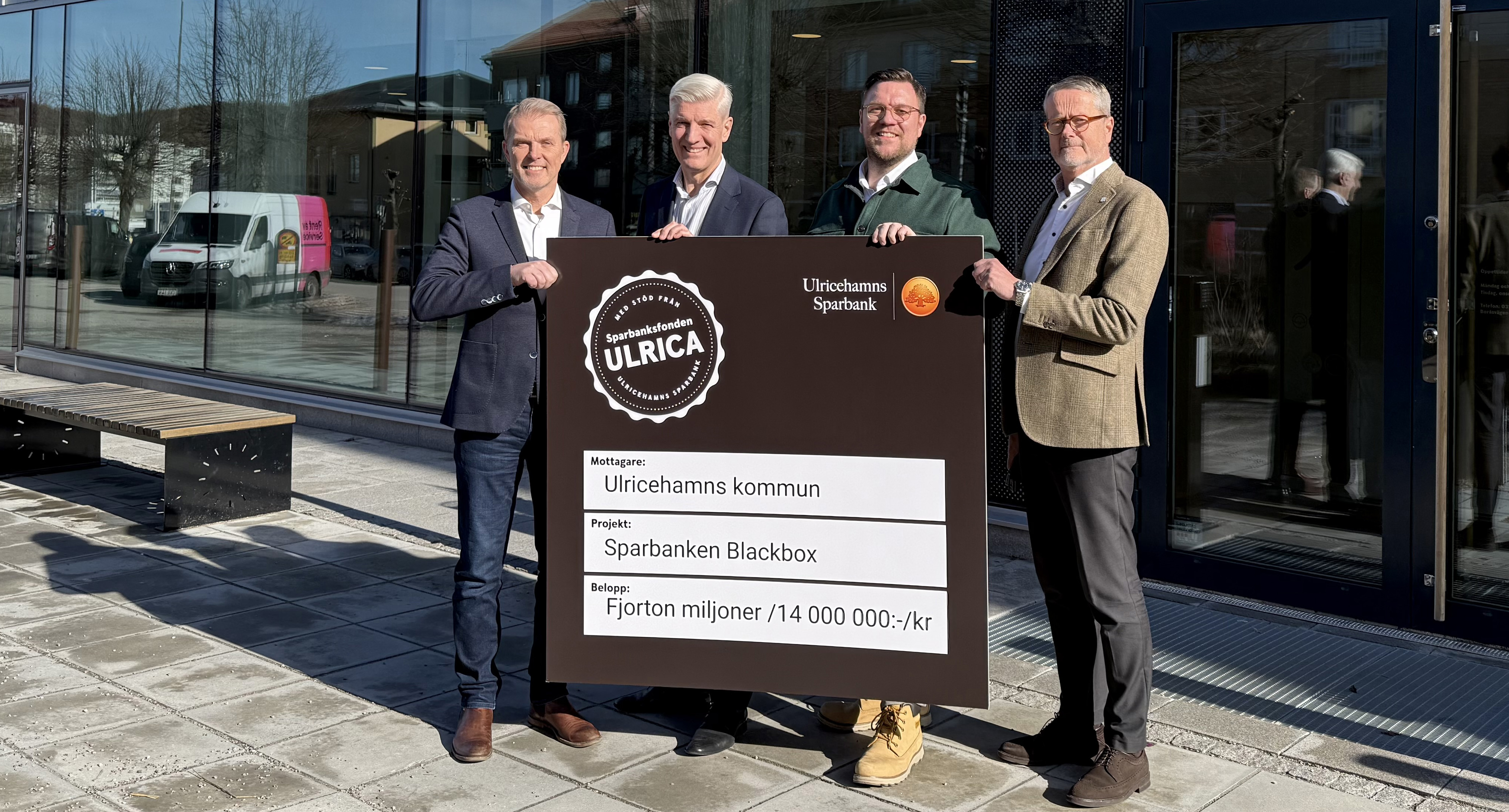 Fyra män står utanför en bankentre, håller i en stort gåvokort , ler och tittar in i kameran